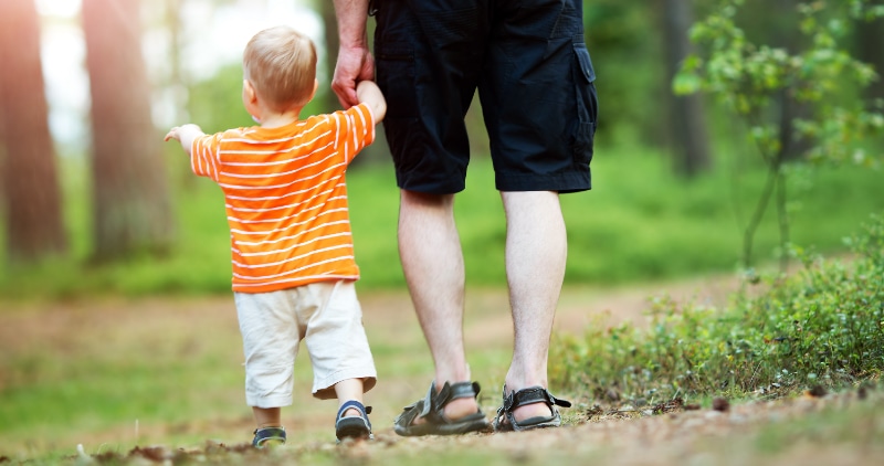 A toddler walks with his parent, holding hands