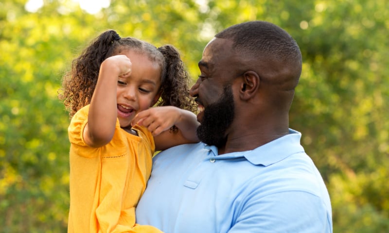 A young Black girl shows her father her bicep